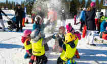 Barn sitter i ring och kastar snö i gula skidskolevästar